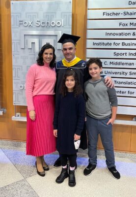 Man in cap and gown stands with a woman and two children in front of a series of plaques