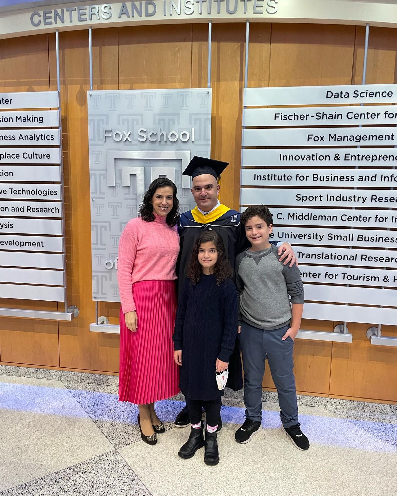 Man in cap and gown stands with a woman and two children in front of a series of plaques