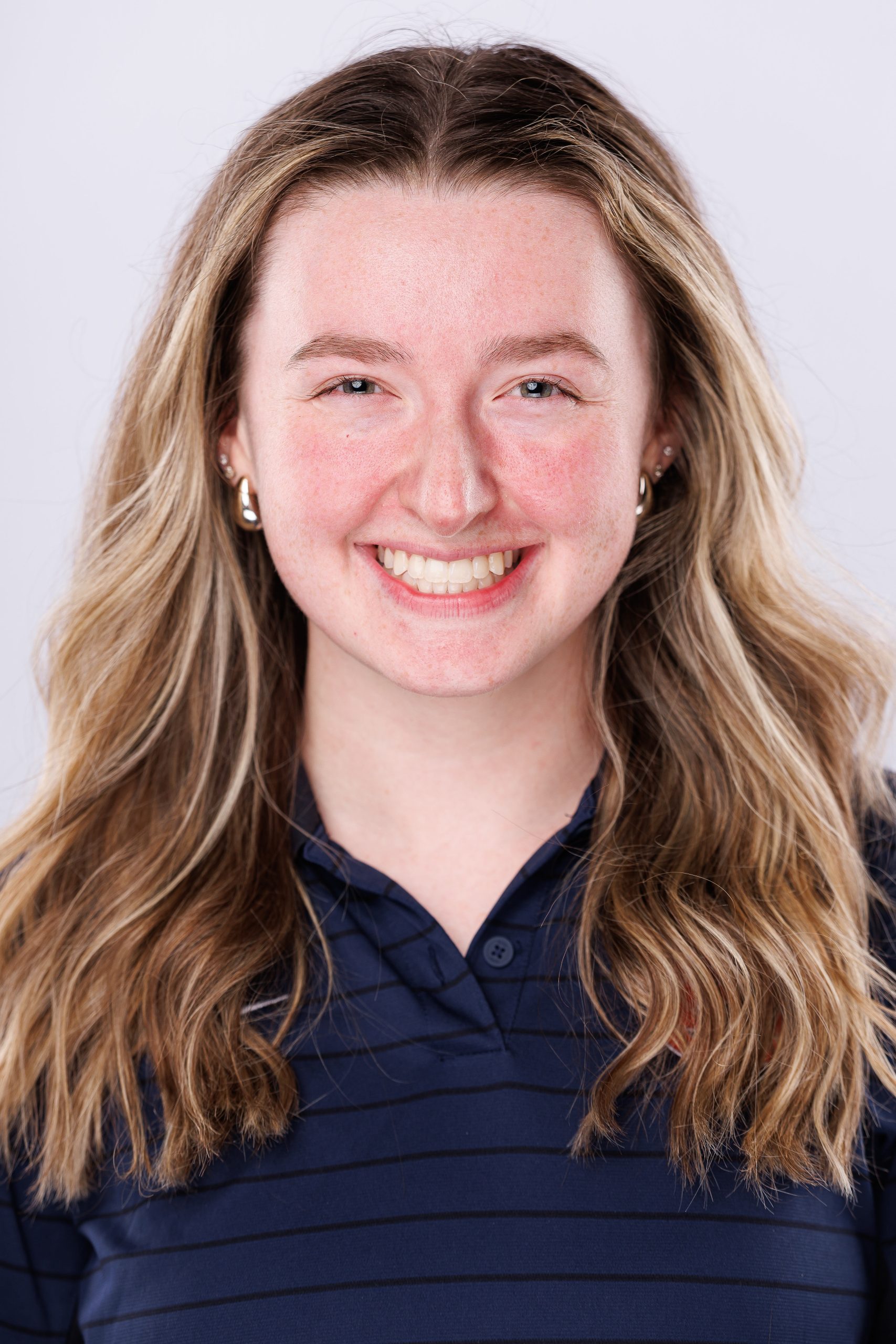 A smiling person with long, wavy blonde hair wearing a dark blue striped polo shirt and small hoop earrings, set against a plain white background.